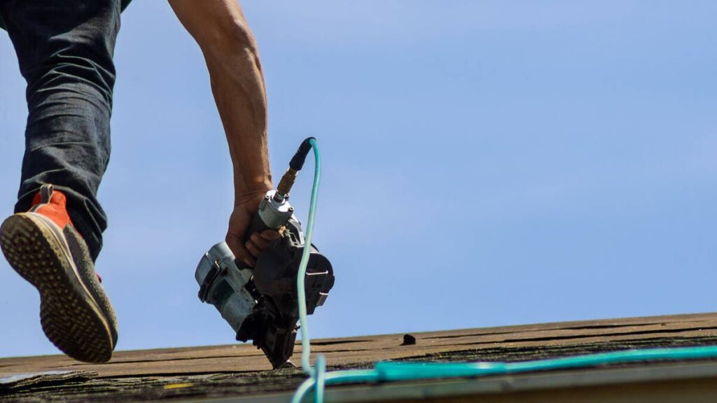 A crew member nailing asphalt shingles with an air compressor nail gun